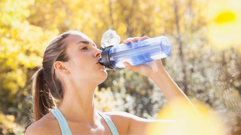 A woman drinking water with fresh fruits and a glass bottle, symbolizing healthy hydration habits for energy, clear skin, and focus.